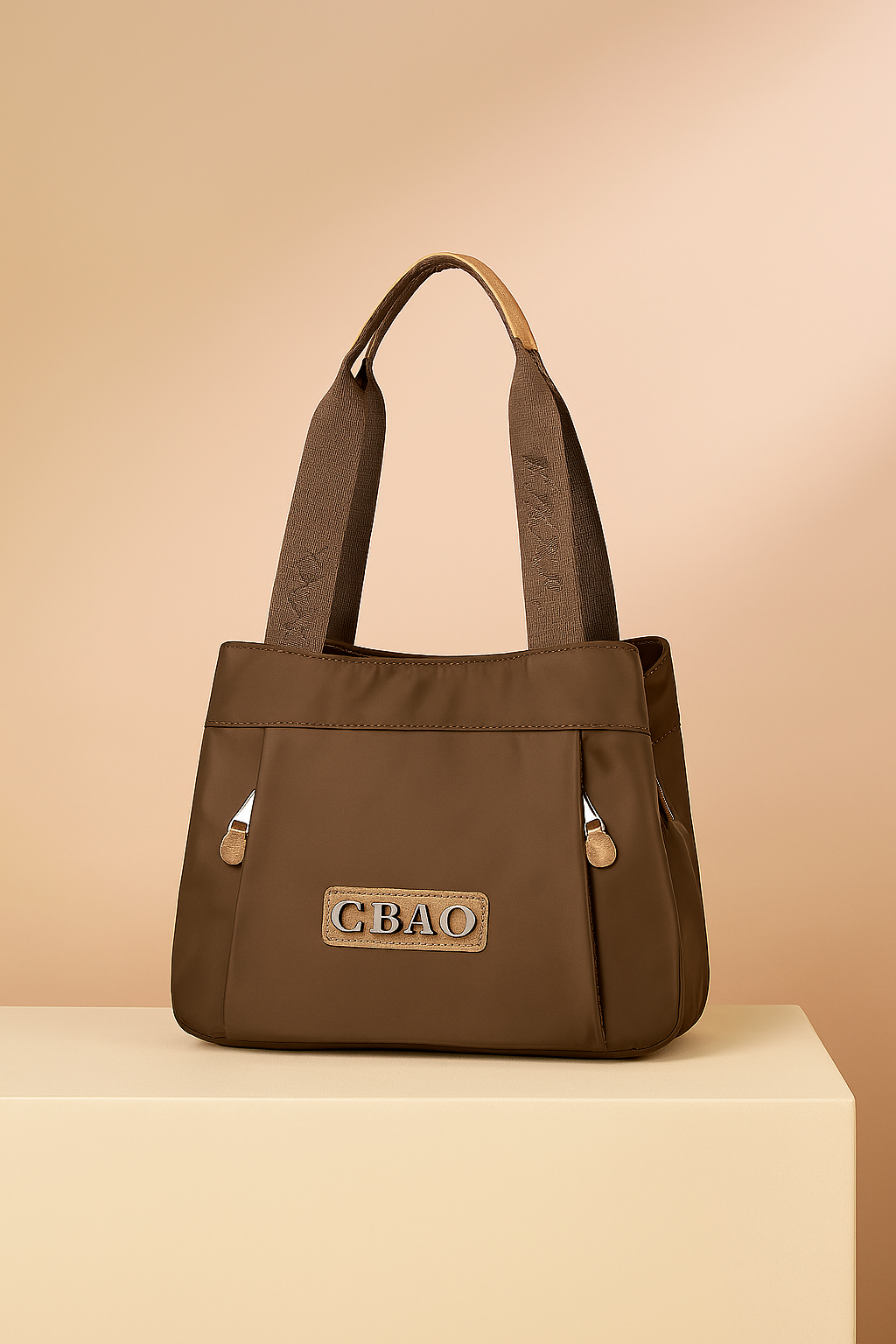 Brown nylon shoulder bag with wide logo straps and front CBAO patch, featuring dual front zip tabs, presented on a neutral platform with a warm sand and pastel pink gradient background.