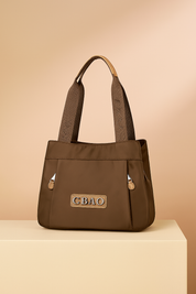Brown nylon shoulder bag with wide logo straps and front CBAO patch, featuring dual front zip tabs, presented on a neutral platform with a warm sand and pastel pink gradient background.