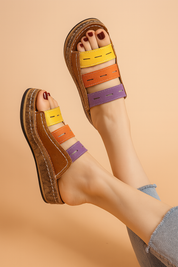 Colorful open-toe wedge sandals featuring three horizontal straps in lavender, orange, and yellow with topstitch details, paired with a textured brown leather base and thick cork platform sole.