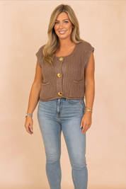 Black knit sleeveless button-front blouse with large gold buttons, styled with light-wash high-rise jeans and nude heeled sandals, photographed in a soft pastel fashion studio background.