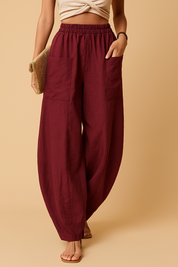 Burgundy wide-leg linen pants with large front patch pockets and elastic waistband, styled with beige sandals and a woven clutch.