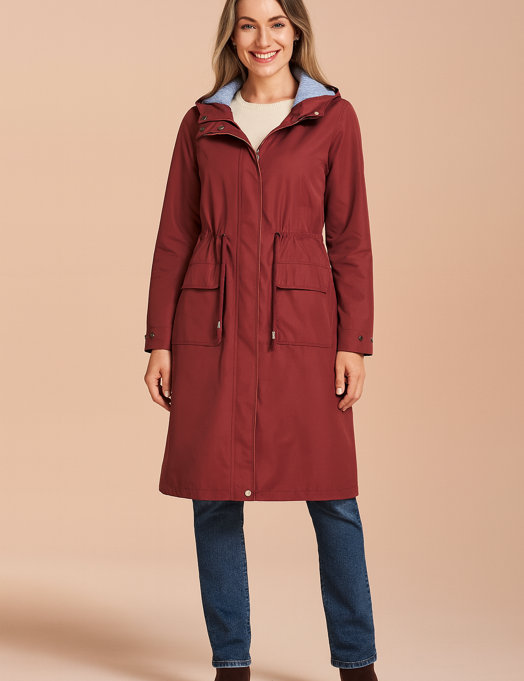 Burgundy knee-length raincoat with drawstring waist, flap pockets, and zip-front closure, layered over a cream knit top, paired with straight-leg blue jeans and brown suede ankle boots.