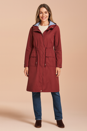 Burgundy knee-length raincoat with drawstring waist, flap pockets, and zip-front closure, layered over a cream knit top, paired with straight-leg blue jeans and brown suede ankle boots.