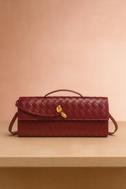 Rectangular burgundy woven leather handbag with a gold knot clasp, top handle, and wrap-around leather strap, displayed on a beige table with a soft blush gradient backdrop.