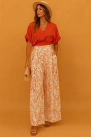 Elegant two-piece set featuring a burnt-orange short-sleeve blouse paired with high-waisted wide-leg trousers in a cream base with orange floral print, accessorized with a straw clutch and matching straw hat for a refined, sunny-day look.