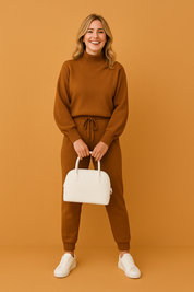 Burnt sienna co-ord set with high-neck pullover and drawstring jogger pants, styled with white sneakers and a structured handbag, captured in a warm sandy studio setting.