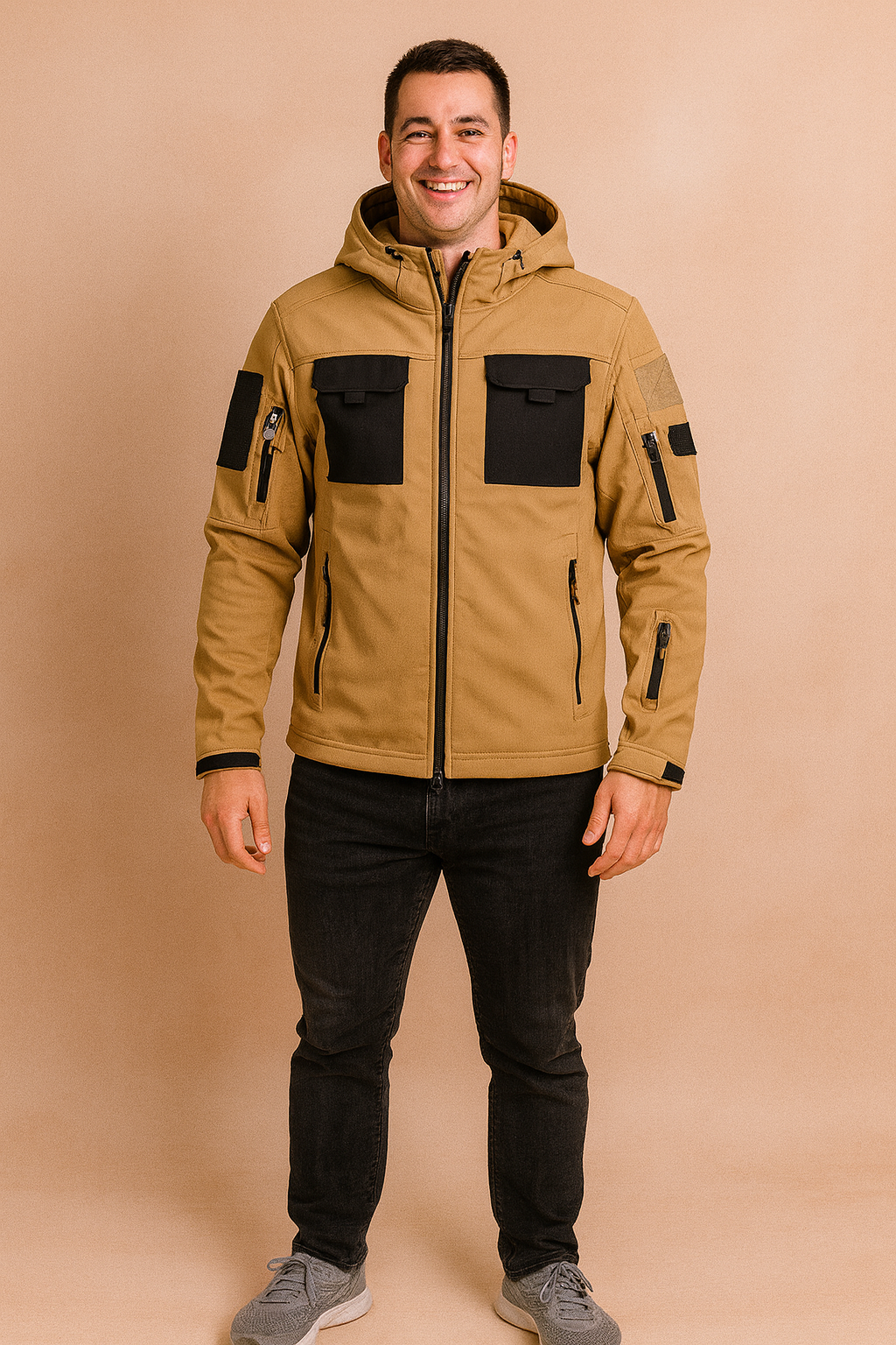 Camel beige tactical softshell jacket with black chest pockets, multiple zippers, and adjustable hood, styled with slim black jeans and grey sneakers.
