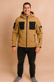 Camel beige tactical softshell jacket with black chest pockets, multiple zippers, and adjustable hood, styled with slim black jeans and grey sneakers.