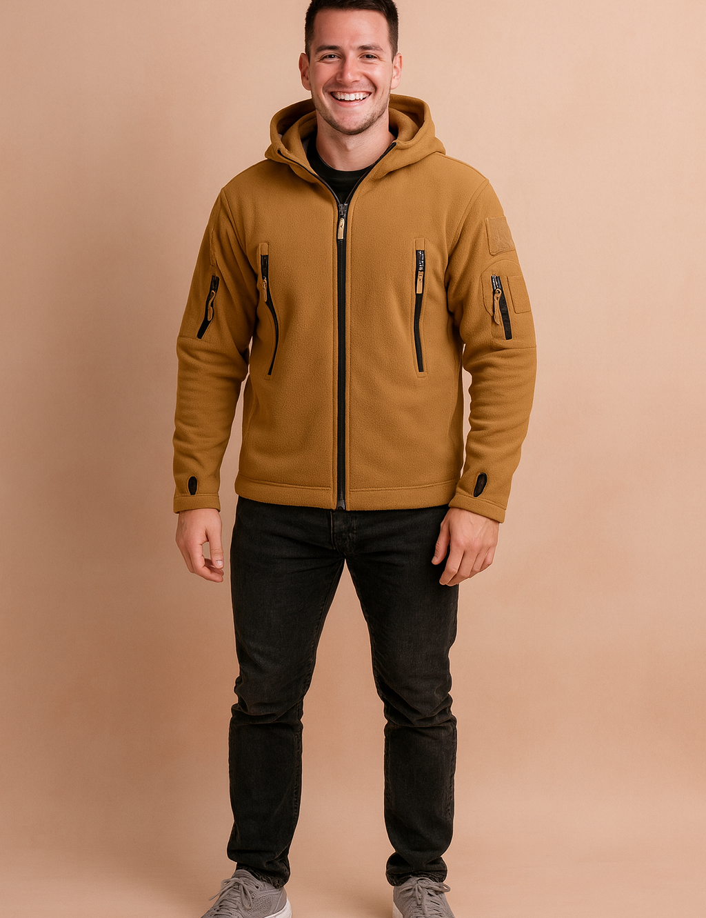 Camel brown softshell jacket with hood, multiple zippered pockets, and thumbhole sleeves, styled with black jeans and grey sneakers.