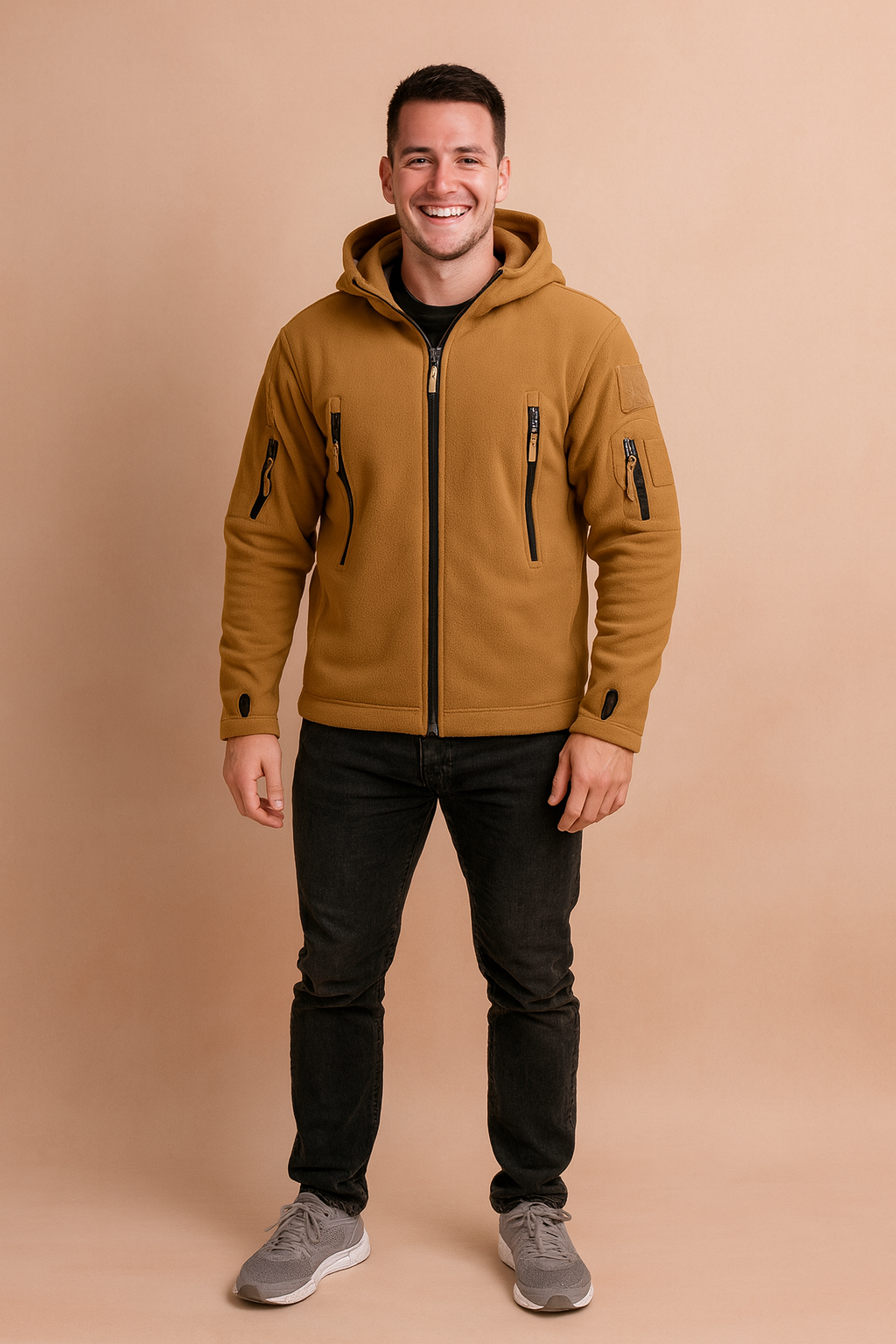 Camel brown softshell jacket with hood, multiple zippered pockets, and thumbhole sleeves, styled with black jeans and grey sneakers.