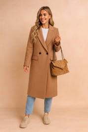 Camel double-breasted wool coat with black buttons, paired with light blue straight-leg jeans, beige chunky sneakers, and a quilted tan chain-strap handbag.