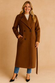 Camel-colored long overcoat with double-breasted button closure, wide lapel collar, belted waist tie, and side pockets, styled over a ribbed white turtleneck sweater and slim blue jeans.