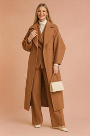 Camel double-breasted long coat with wide lapels, paired with matching wide-leg trousers, a white turtleneck top, pointed-toe heels, and a cream handbag with gold chain strap.