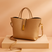 Camel-toned pebbled leather tote bag with a structured trapezoid shape, double top handles, a gold-tone vertical clasp strap, and a long matching shoulder strap, displayed on a neutral podium in soft lighting.