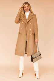 Camel double-breasted wool coat with gold-toned buttons, paired with a white turtleneck, cream trousers, and white ankle boots, accessorized with a beige patterned handbag.
