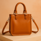 Structured caramel brown leather handbag with double top handles, silver hardware, and an adjustable shoulder strap, displayed on a neutral platform under soft studio lighting.