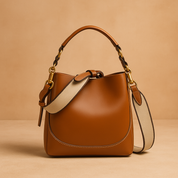  Caramel brown leather saddle bag with curved front panel, gold hardware, and cream canvas strap, placed on a warm sand and pastel pink gradient backdrop with soft studio lighting.