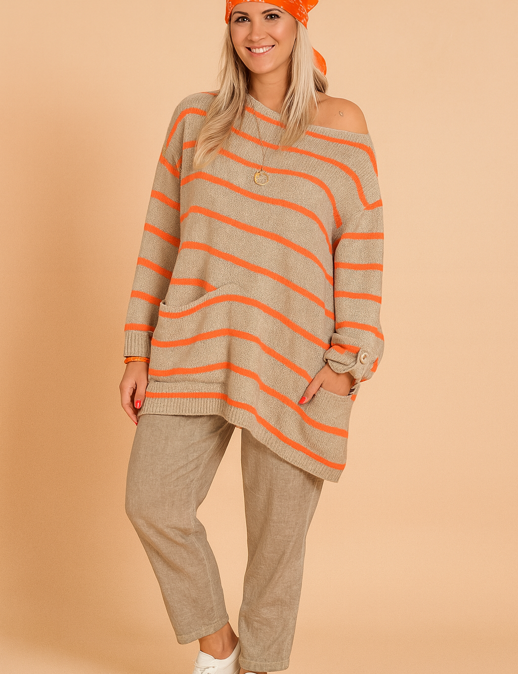 Oversized beige knit sweater with bright orange horizontal stripes, asymmetrical hem, front pocket, and button-cuff long sleeves, paired with loose-fit taupe linen trousers and white sneakers.