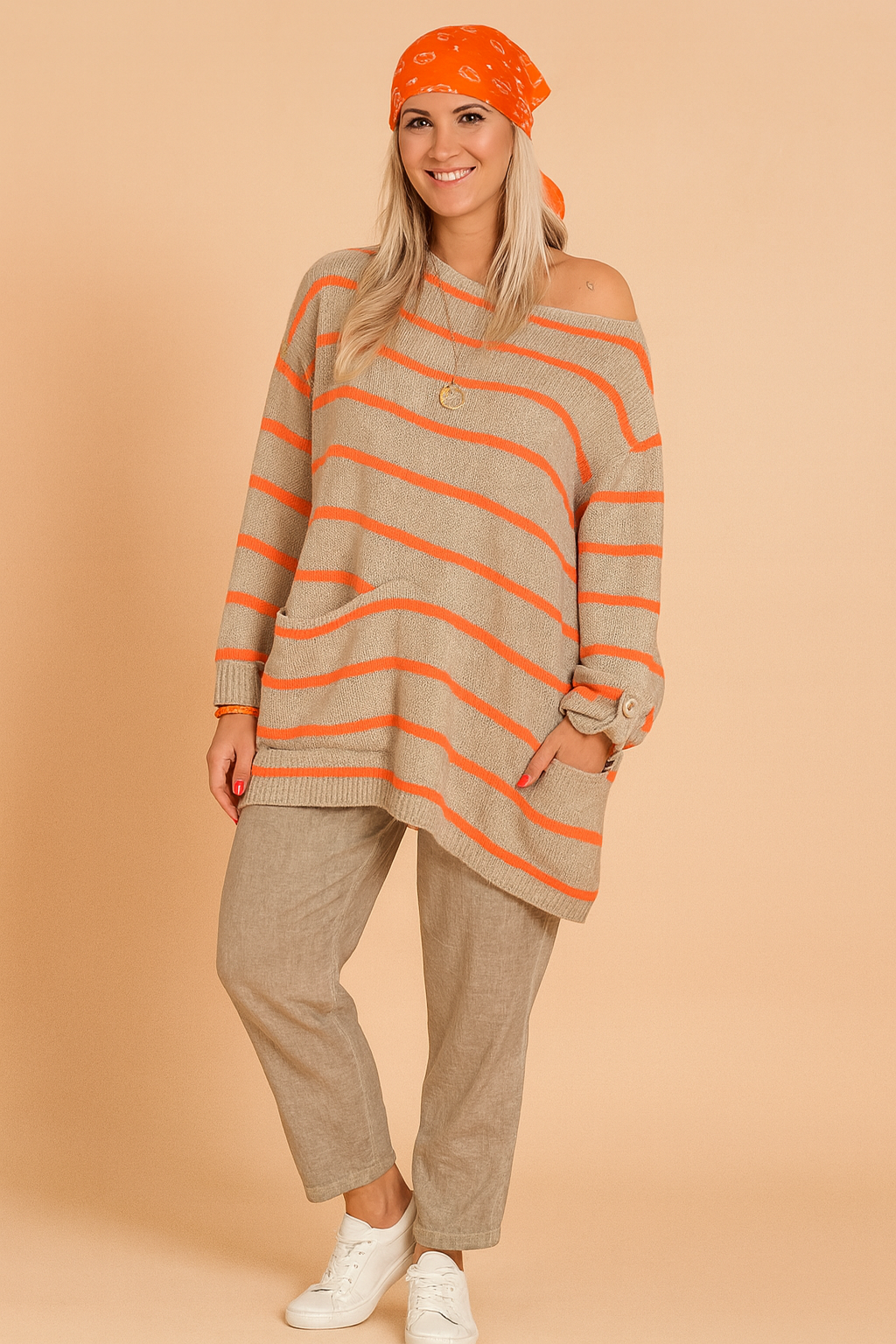 Oversized beige knit sweater with bright orange horizontal stripes, asymmetrical hem, front pocket, and button-cuff long sleeves, paired with loose-fit taupe linen trousers and white sneakers.