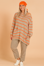 Oversized beige knit sweater with bright orange horizontal stripes, asymmetrical hem, front pocket, and button-cuff long sleeves, paired with loose-fit taupe linen trousers and white sneakers.