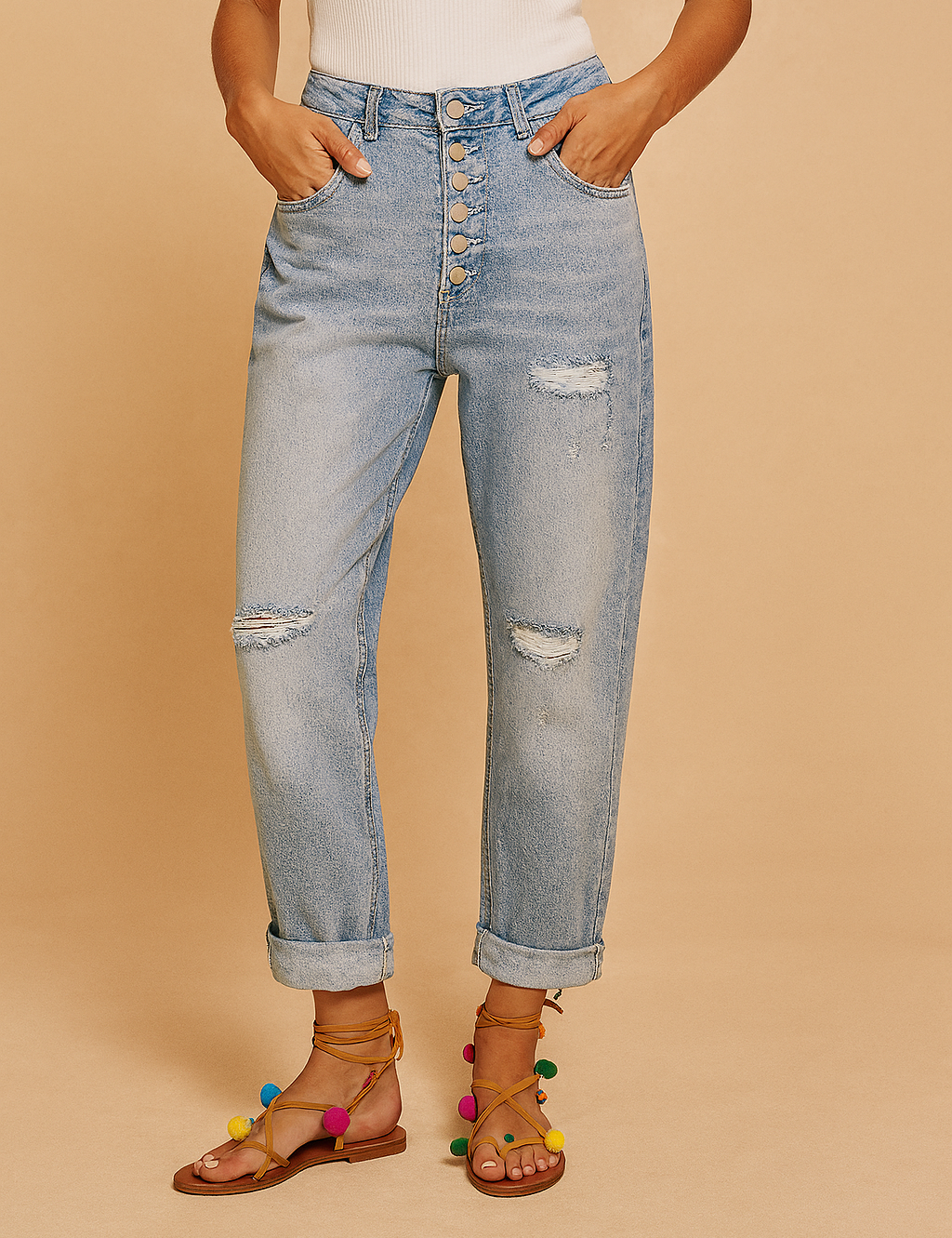 High-waisted light wash denim jeans with six metallic front buttons, distressed patches on the thighs and knees, cuffed hems, and styled with multicolored pom-pom tie-up sandals.
