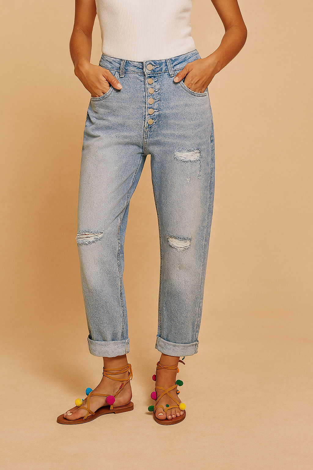 High-waisted light wash denim jeans with six metallic front buttons, distressed patches on the thighs and knees, cuffed hems, and styled with multicolored pom-pom tie-up sandals.