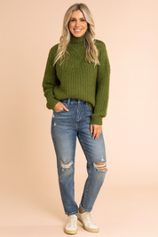 Chunky olive green cable-knit turtleneck sweater paired with distressed blue high-waisted jeans and white low-top sneakers.