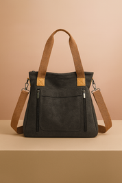 Charcoal grey canvas tote bag with brown shoulder straps, silver zipper details, and front pockets, displayed on a beige platform with a warm sand and pastel pink gradient background.