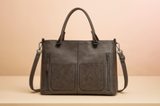 Dark grey-brown leather tote handbag with dual top handles, detachable shoulder strap, two front patch pockets, and vertical zipper compartments, presented on a beige table with a soft pastel gradient background.