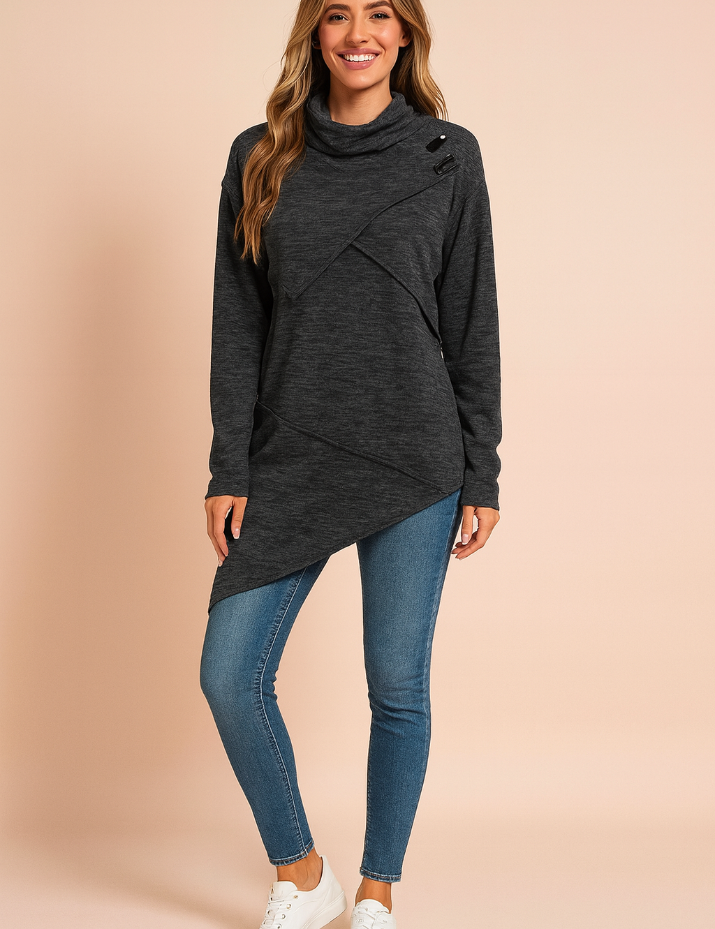 Dark charcoal grey asymmetrical tunic sweater with diagonal wrap design, high cowl neck, and button detail on the shoulder, paired with fitted mid-wash skinny jeans and white lace-up sneakers.