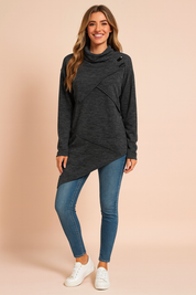 Dark charcoal grey asymmetrical tunic sweater with diagonal wrap design, high cowl neck, and button detail on the shoulder, paired with fitted mid-wash skinny jeans and white lace-up sneakers.