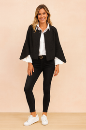 Black wool-blend cape jacket with front button closure, layered over a crisp white button-up shirt, paired with fitted black skinny jeans and white low-top sneakers.