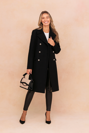 Elegant black double-breasted wool coat with silver-tone buttons, paired with sleek black leather leggings and black pointed-toe heels, complemented by a two-tone beige and black handbag.