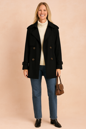 Black double-breasted trench coat with wide lapels, buttoned cuffs, and brown buttons, layered over a cream turtleneck sweater, paired with straight-leg blue jeans and black ankle boots, accessorized with a small brown leather handbag.