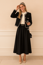 Elegant black tweed jacket with pearl-trimmed edges and gold button detailing, styled over a white ruffled blouse and paired with a flowing black skirt and nude heels.
