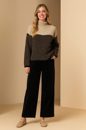 Beige and black color-blocked high-neck sweater with a bold chevron pattern on the lower half and sleeves, paired with black straight-leg trousers and beige lace-up sneakers.