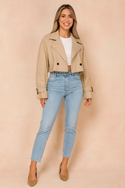 Cropped beige trench coat with black button details, paired with a fitted white crop top, high-waisted light wash jeans, and tan pointed-toe heels.