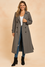 Long double-breasted gray trench coat with wide lapels, matching belt detail on cuffs, paired with a white scoop-neck top, slim-fit distressed blue jeans, and black suede ankle boots.