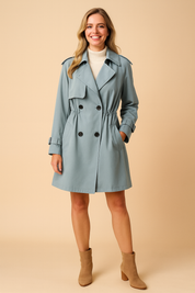  Light blue double-breasted trench coat with cinched waist, epaulettes, buckled sleeve straps, and black buttons, paired with a white turtleneck sweater and beige ankle boots.