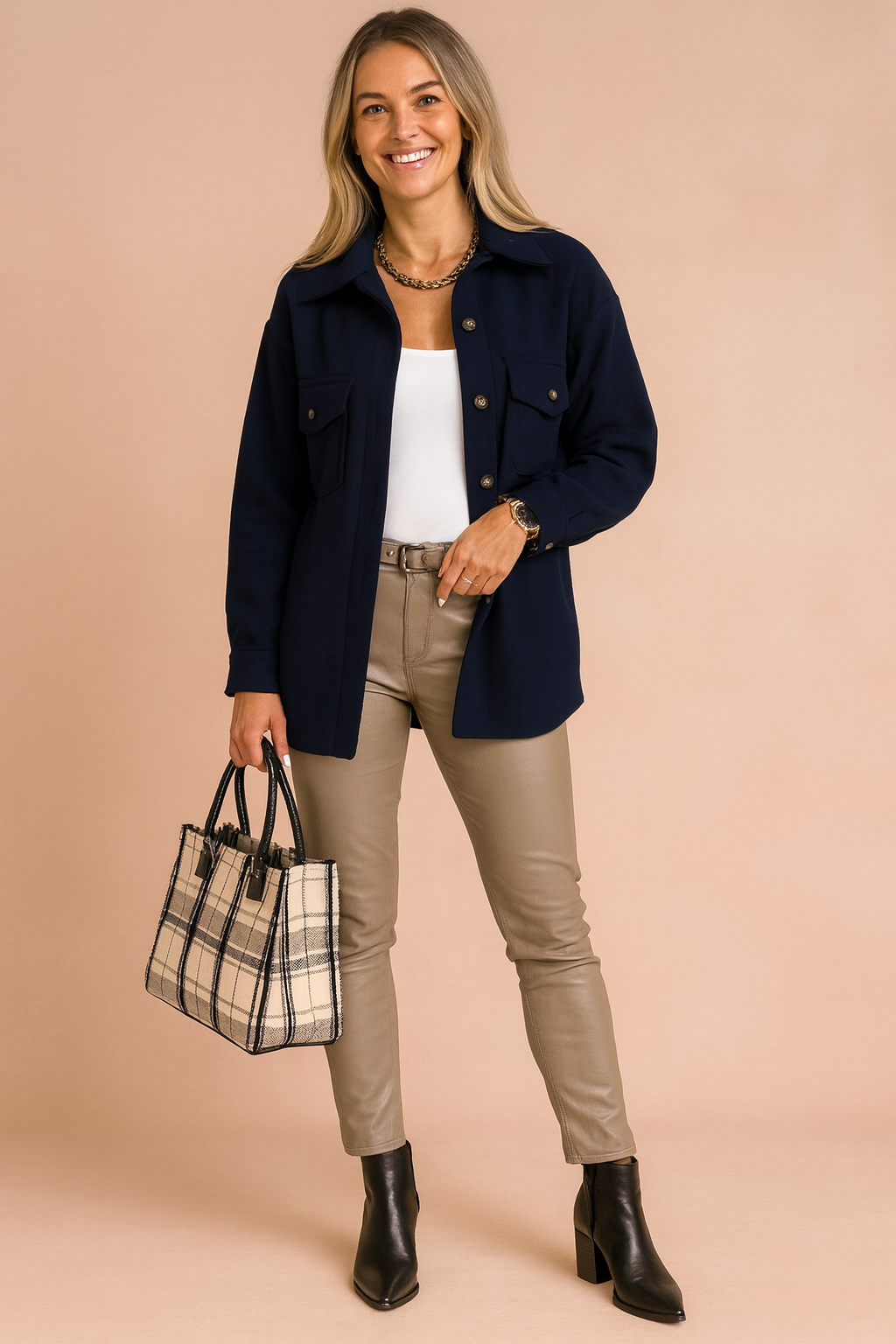 Navy blue button-up shirt jacket with front flap pockets, paired with a white tank top, beige fitted leather-look pants, black ankle boots, and a plaid structured handbag.