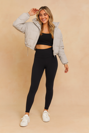 Light gray cropped puffer jacket with front zipper and snap buttons, paired with a black sports bra and high-waisted black leggings, styled with classic white lace-up sneakers.