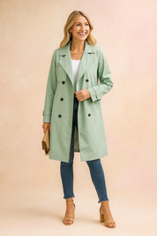 Light sage green double-breasted trench coat with black buttons, paired with a white top, dark blue skinny jeans, tan block-heeled sandals, and a beige clutch bag.