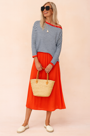 Navy and white striped off-shoulder sweater with red trim, paired with a vibrant red midi skirt, natural woven tote bag, and cream espadrille flats.