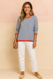 Navy and white horizontal striped sweater with bright red ribbed trim at the neckline, cuffs, and hem, paired with straight-leg white trousers and beige espadrilles.