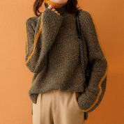 Chunky ribbed knit turtleneck sweater in mustard and charcoal with beige trousers, styled with black leather bag against an orange sandy studio wall.