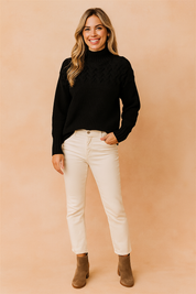 Black cable-knit high-neck sweater with intricate textured yoke design, paired with slim-fit cream jeans and taupe suede ankle boots.