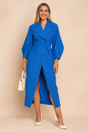 Elegant cobalt blue trench coat dress with balloon sleeves, double-breasted button closure, and thigh-high slit, paired with white pointed-toe heels and an off-white handbag.