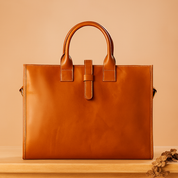 Rectangular cognac leather tote bag with structured top handles and front strap detail, displayed on a wooden platform against a warm sand and pastel pink gradient background.