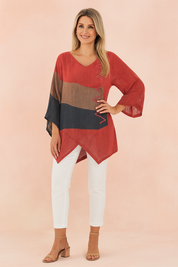 Asymmetrical red tunic blouse with brown and navy color-blocked stripes, featuring zigzag stitched accents and loose 3/4 sleeves, paired with white fitted pants and nude sandals.