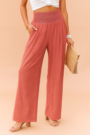 High-waisted coral red wide-leg pants with a smocked waistband and soft pleats, styled with beige open-toe block heel sandals and a straw clutch.